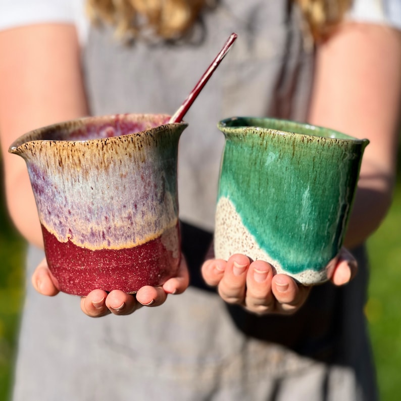 May include: Two handmade ceramic mugs. One mug is red, purple, and tan with a spout and a red stirring stick. The other mug is green and tan. Both mugs have a rustic, textured appearance and are held by a person.