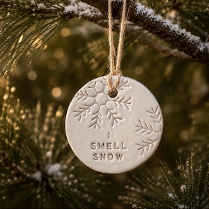 May include: A round, off-white ceramic ornament featuring an embossed snowflake design and the stamped text "I SMELL SNOW" in capital letters. The ornament is suspended by a thin, natural-colored jute twine loop through a hole at the top.