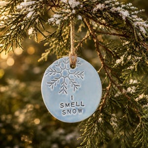May include: A round, light blue ceramic ornament featuring an embossed snowflake design and the text I SMELL SNOW stamped in black lettering. The ornament hangs from a natural jute twine loop, creating a simple and clear decorative piece.