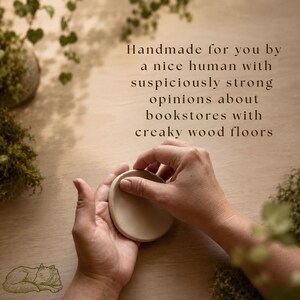 May include: A small, round, beige clay disc held between two hands. The text reads: Handmade for you by a nice human with suspiciously strong opinions about bookstores with creaky wood floors. A small line drawing of a sleeping cat is in the corner.