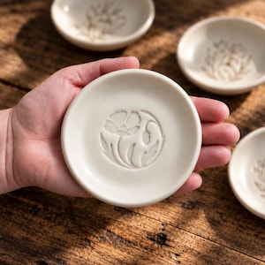 May include: A small, round, off-white ceramic dish featuring a debossed floral design in the center. The dish has a smooth, matte finish and a slightly raised rim. Several similar dishes with matching floral patterns are visible in the background.