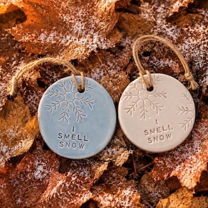 May include: Two circular ceramic ornaments with twine hangers. One is light blue and the other is off-white. Both feature an embossed snowflake design and the text "I SMELL SNOW" stamped in the center. The ornaments have a smooth, glazed finish.