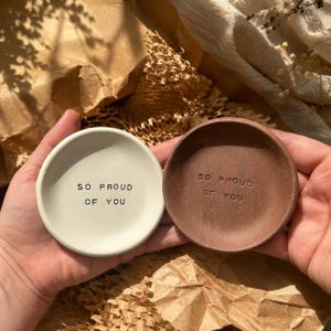 May include: Two circular trinket dishes, one in a smooth off-white finish and the other in a textured dark brown clay finish. Both dishes feature the stamped black text "SO PROUD OF YOU" centered on the interior base of each individual bowl.