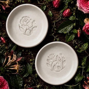May include: Two circular white ceramic dishes with raised relief designs. The top dish features a single rose blossom with leaves, while the bottom dish displays a stylized floral arrangement with multiple petals and stems centered on the base.