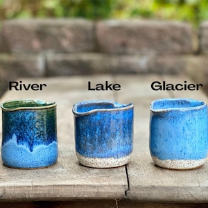 May include: Three handmade ceramic cups with unique blue glaze finishes. Each cup is labeled with a name: "River", "Lake", and "Glacier". The cups have a rustic, textured appearance and are set on a wooden surface.