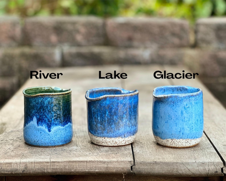 May include: Three handmade ceramic cups with unique blue glaze finishes. Each cup is labeled with a name: "River", "Lake", and "Glacier". The cups have a rustic, textured appearance and are set on a wooden surface.