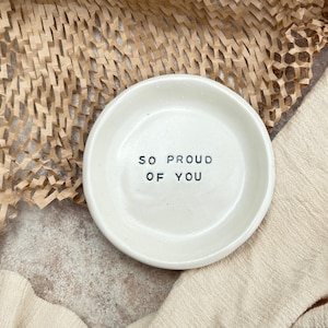 May include: White ceramic trinket dish with the text "So Proud Of You" in black lettering.