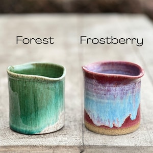 May include: Two handmade ceramic cups, one in forest green and the other in frostberry. The forest green cup has a glossy finish, while the frostberry cup features a blend of red, blue, and purple hues. Both are cylindrical with a slightly curved rim.