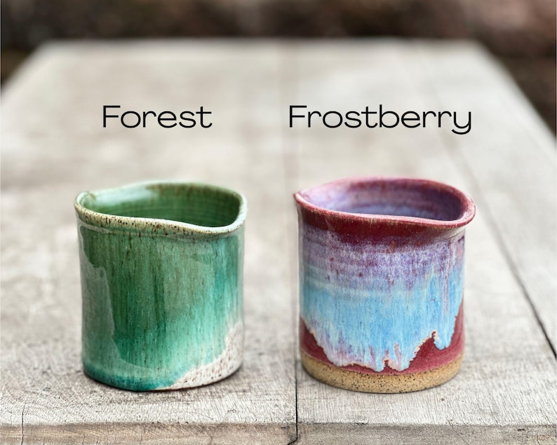 May include: Two handmade ceramic cups, one in forest green and the other in frostberry. The forest green cup has a glossy finish, while the frostberry cup features a blend of red, blue, and purple hues. Both are cylindrical with a slightly curved rim.