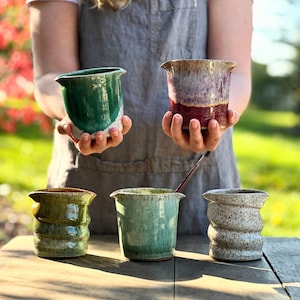 May include: A collection of handmade ceramic pottery. The image features four unique pieces: a green and white vase, a purple and red vase, a green vase with a ribbed design, a light green vase, and a speckled vase with a ribbed design. The pottery is displayed on a wooden surface.
