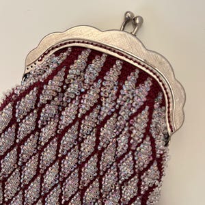 Può includere: Primo piano di una borsetta vintage con perline. La borsetta presenta una base in tessuto bordeaux scuro ricoperta da un motivo a diamante di perline iridescenti. La cornice è color argento con un bordo smerlato e una chiusura a scatto.