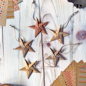 May include: Five origami stars made from patterned paper with twine hangers. The stars are in various colors and patterns, including red, brown, and white. The stars are on a light wood background.