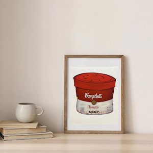 May include: A framed print of a Campbell's Tomato Soup can. The artwork features a red can lid with a white and gold label that reads "Tomato Soup." The print is in a wooden frame and is displayed on a light-colored surface.