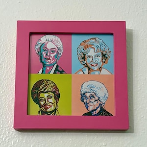 May include: A framed pop art print featuring four portraits of women, each in a different quadrant. The portraits are set against pink, blue, yellow, and orange backgrounds, framed in a bright pink border. The artwork has a vibrant, colorful style.
