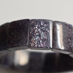 Puede incluir: Un brazalete negro hecho de cuentas cuadradas con purpurina iridiscente incrustada en la superficie.