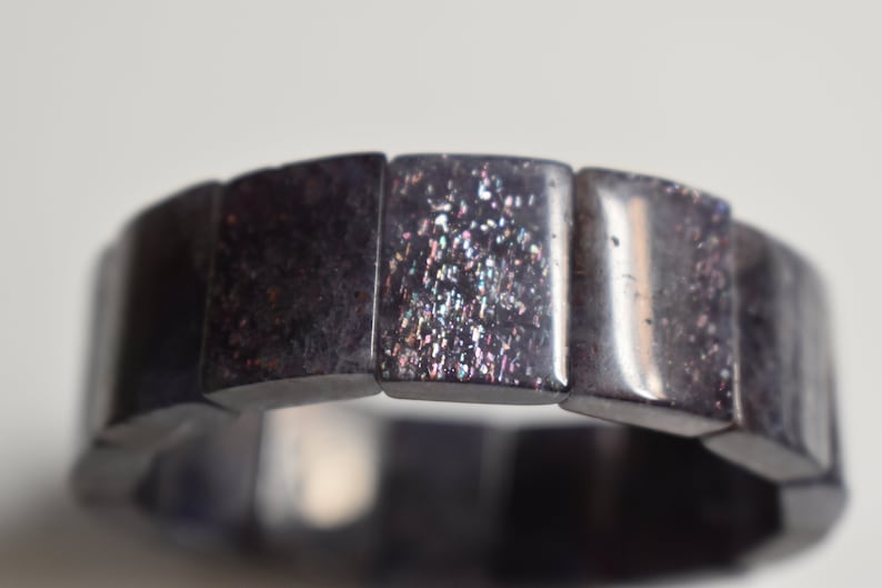 Puede incluir: Un brazalete negro hecho de cuentas cuadradas con purpurina iridiscente incrustada en la superficie.