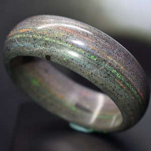 May include: A gray and iridescent ring with a smooth, polished surface. The ring has a subtle sparkle and a unique, textured look.