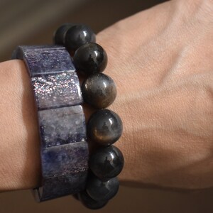Puede incluir: Dos pulseras en una mu&ntilde;eca. Una pulsera est&aacute; hecha de cuentas cuadradas de color azul oscuro con un acabado brillante. La otra pulsera est&aacute; hecha de cuentas redondas de color gris oscuro con un brillo dorado.