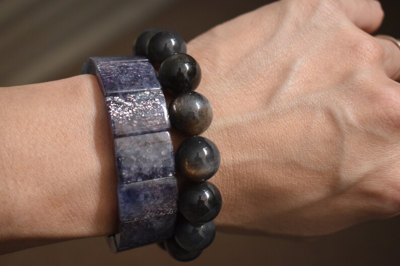 Puede incluir: Dos pulseras en una mu&ntilde;eca. Una pulsera est&aacute; hecha de cuentas cuadradas de color azul oscuro con un acabado brillante. La otra pulsera est&aacute; hecha de cuentas redondas de color gris oscuro con un brillo dorado.