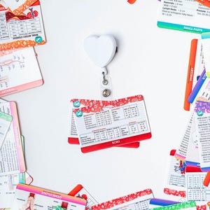 May include: A white heart-shaped retractable badge holder with a variety of colorful medical reference cards scattered around it. The cards contain medical terminology, diagrams, and measurements.
