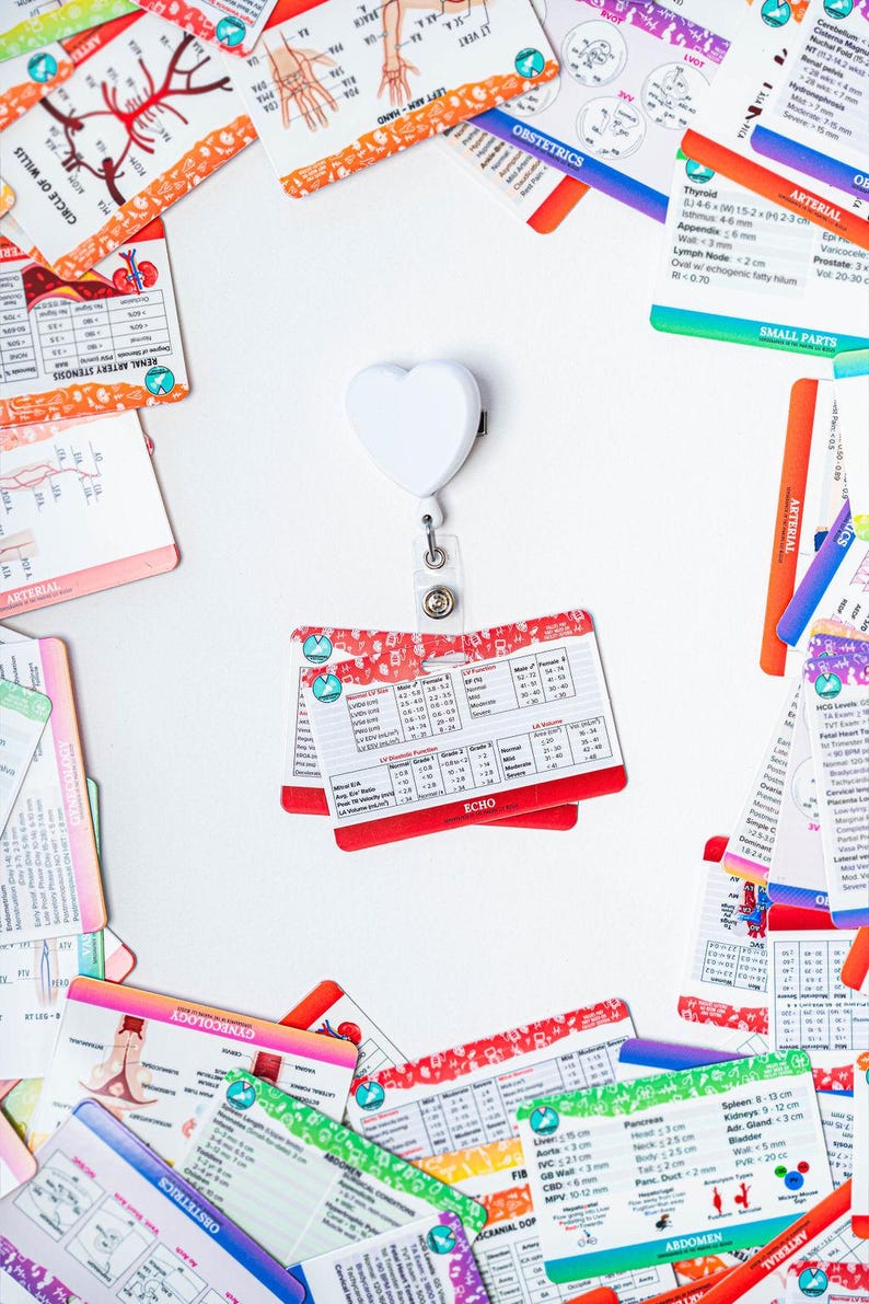 May include: A white heart-shaped retractable badge holder with a variety of colorful medical reference cards scattered around it. The cards contain medical terminology, diagrams, and measurements.