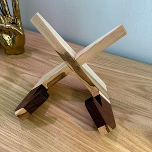 May include: A wooden stand with a cross-shaped design. The stand is made of two different types of wood, one light and one dark. The stand is designed to hold a small object, such as a phone or a book.