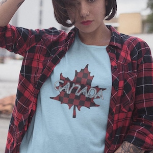 Puede incluir: Una camiseta azul claro con un diseño de hoja de arce a cuadros rojos y negros y la palabra "CANADA" en letras plateadas. La persona lleva una camisa de franela a cuadros rojos y negros.