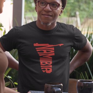 Peut inclure: Un t-shirt noir avec un motif rouge de la carte de la Namibie et le mot "Namibie" en police verticale. Le t-shirt est porté par une personne avec des lunettes et un chapeau.