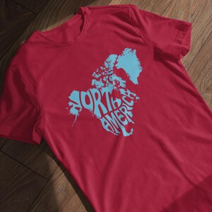 May include: A red t-shirt featuring a light blue graphic of the North American continent with the words "NORTH AMERICA" in a stylized font. The shirt is made of a soft material and is displayed on a wooden surface.