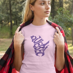 Könnte beinhalten: Hellrosa T-Shirt mit einem lila Grafik der Provinz Quebec und dem Wort "QUEBEC" in einer stilisierten Schriftart. Die Person trägt eine rot-schwarz karierte Decke über den Schultern.