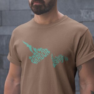 May include: A brown t-shirt with a teal graphic design featuring the word "Labrador" and a stylized map outline. The shirt has rolled-up sleeves and a crew neck.