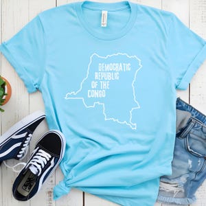 Puede incluir: Una camiseta azul claro con un contorno blanco de la República Democrática del Congo y el texto "República Democrática del Congo" impreso en ella.
