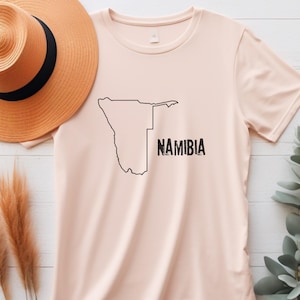 Peut inclure: Un t-shirt rose pâle avec un contour noir du pays de Namibie et le mot "NAMIBIA" imprimé en noir.