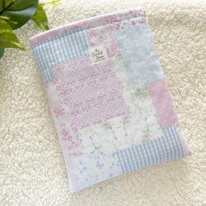 May include: A patchwork fabric case featuring various pastel patterns, including floral prints, gingham, and solid colors. The case has a label that reads "The Crochet Fiends". The case is designed to hold a book or tablet.