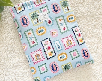 Op de afbeelding: Een lichtblauwe stoffen boekenhoes met een herhalend patroon van ingelijste tropische afbeeldingen. De frames bevatten afbeeldingen van palmbomen, schelpen, vissen en bloemen. De tekst "The Comfort Finds" staat op een klein wit label.