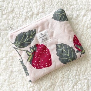 May include: A quilted zippered pouch in light pink featuring a pattern of red strawberries and dark green leaves. A white rectangular fabric label is sewn onto the front with the black text The Crochet Ficus printed on it.