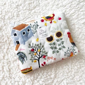 May include: A quilted fabric zipper pouch featuring a white background with a colorful print of a blue barn, sunflowers, fruit trees, chickens, and small red apples. A white square label in the center displays the text The Crochet Ficus.