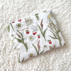 May include: A white quilted zippered pouch featuring a repeating pattern of small brown mice, red strawberries, green stems, and white flowers. A small square fabric label in the center displays the text The Crochet Ficus.