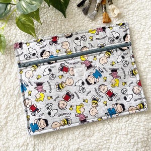 Può includere: Una custodia rettangolare in tessuto grigio con un motivo ripetuto di personaggi Peanuts, tra cui Snoopy, Charlie Brown, Lucy, Linus, Sally e Woodstock. La custodia presenta una chiusura a zip grigia e una piccola etichetta bianca The Crochet Fix.
