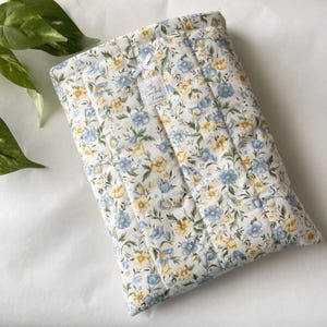May include: A quilted floral laptop sleeve featuring a pattern of blue and yellow flowers on a white background. The sleeve has a rectangular shape with vertical stitching and a small tag that reads "The Crochet Ficcus".