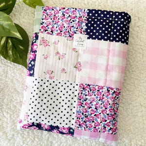 Op de afbeelding: Een patchwork item met diverse patronen, waaronder bloemenprints, stippen en ruitjes. Het kleurenpalet omvat roze, wit en marineblauw. Het item heeft een label met de tekst "The Crochet Ficas".