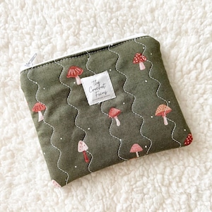 May include: A rectangular olive green zippered pouch featuring a repeating pattern of small red and white mushrooms. The fabric has white wavy quilted stitching and a white fabric label in the center that reads The Crochet Ficus.