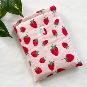 May include: A pink quilted sleeve with a strawberry pattern. The sleeve features a repeating design of red strawberries with green stems and leaves on a light pink background with white polka dots. The label reads "The Crochet Ficup".