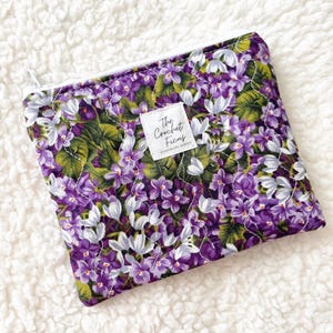 May include: A rectangular zippered pouch featuring a vibrant floral pattern of purple and white violets with green leaves. The fabric has a quilted texture with white stitching. A white rectangular label reads The Crochet Ficus.