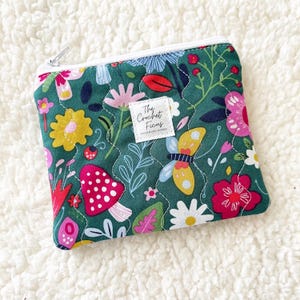 May include: A rectangular zippered pouch with a dark green background featuring a colorful pattern of pink, yellow, and red flowers, mushrooms, and a butterfly. A white square label in the center displays the text The Crochet Ficus.