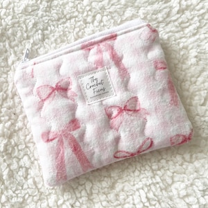 May include: A quilted zippered pouch featuring a white and light pink gingham pattern with repeating pink bow illustrations. A small rectangular white fabric label is sewn on the front with the black text The Crochet Ficus.