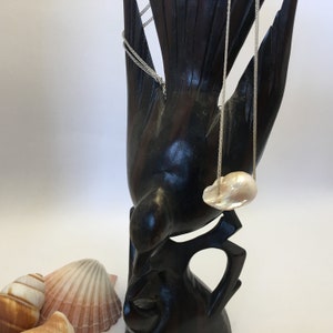 May include: A white baroque pearl pendant necklace hanging from a silver chain on a dark wooden bird sculpture.