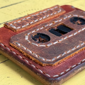 May include: A brown leather wallet with a stitched design resembling a vintage cassette tape. The wallet has a flap closure and white stitching.