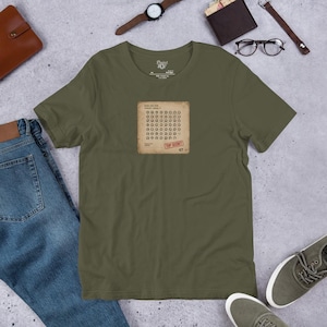 Può includere: Una t-shirt a maniche corte color verde oliva con una grafica rettangolare sul petto. La grafica ricorda un documento invecchiato con una griglia di piccole icone circolari, testo e un timbro rosso nell'angolo in basso a destra con la scritta TOP SECRET 47.
