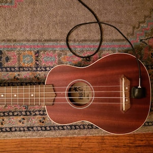 May include: A brown ukulele with a black pickup and a black cable attached to it. The ukulele is lying on a patterned rug.
